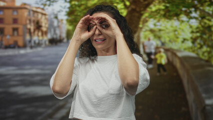 Woman in white cropped tee makes binocular gesture with hands over eyes on sunlit urban street; curiosity.