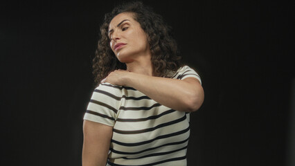 Young hispanic woman in striped shirt with curly hair holds shoulder in black studio; discomfort tension strain pain.