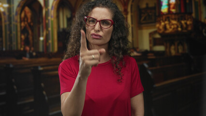 Hispanic woman wearing red shirt and glasses points finger forward inside church building; criticism.