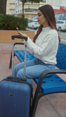 Young woman sitting outdoors on a street bench with a suitcase, using a smartphone while waiting or traveling in an urban setting.