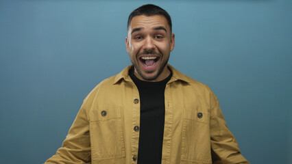 Young hispanic man in mustard jacket covers eyes with hands against blue studio wall in moody pose;...