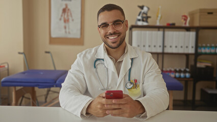 Man doctor looks at red smartphone while seated at a desk in clinic, stethoscope draped over his shoulders; professionalism.