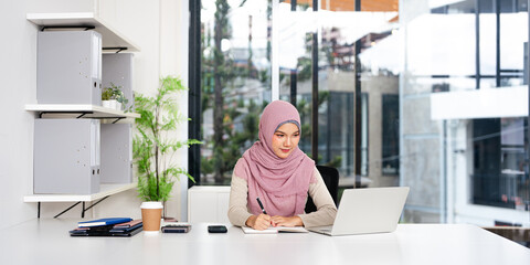 Confident Muslim business woman in hijab, writer  using laptop computer, taking notes, working...