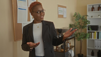 Woman in brown blazer presenting with open palm gesture and glasses in building lobby; business...