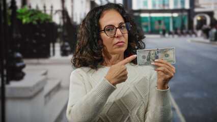 Middle-aged woman in city street pointing at a hundred dollar bill with a thoughtful expression,...