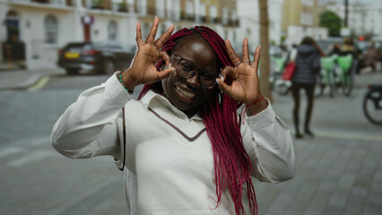 Woman on city street making ok gesture with fingers while smiling, wearing glasses and white top,...