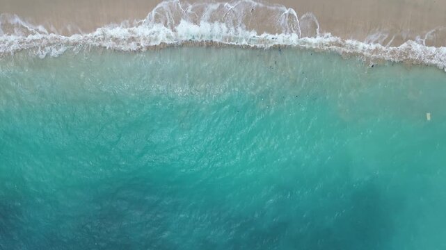 Drone shot of turquoise ocean waves breaking on sandy shore, creating natural abstract seascape with foam and texture.