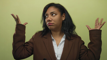 African american woman shrug hands and lift palms wearing brown blazer in studio against light green wall; frustration.