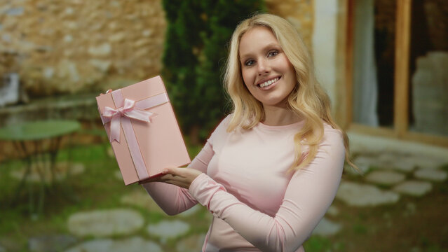 Young woman holding pink gift in outdoor restaurant setting with tables and stone path, smiling joyfully.