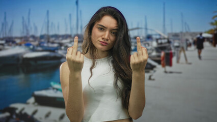 Young hispanic woman shows middle finger to docked boats on a sunlit street walkway by the harbor;...