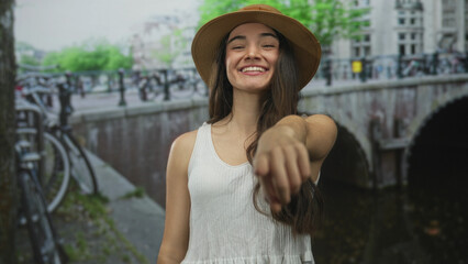 Fototapeta premium Hispanic teenager woman in straw hat pointing finger on city street canal bridge walkway; friendly invitation.