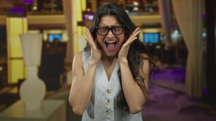 Young woman wearing glasses and striped top presses both hands against cheeks and grimaces in hotel...