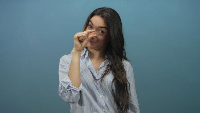 Young hispanic woman pinching fingers to show small size while smiling in blue studio setting; playfulness.