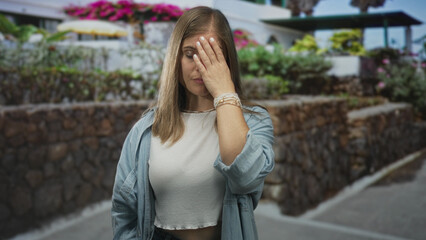 Fototapeta premium Young blonde woman hand raised in bright daylight against stone building by blooming plants on street; confusion.