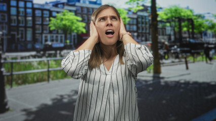 Woman in striped shirt covers ears on busy street beside canal railings under bright sunny sky;...