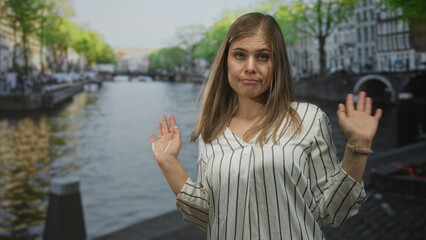 Woman wearing striped blouse raises hands beside canal water on amsterdam street with busy city buildings; indifference.