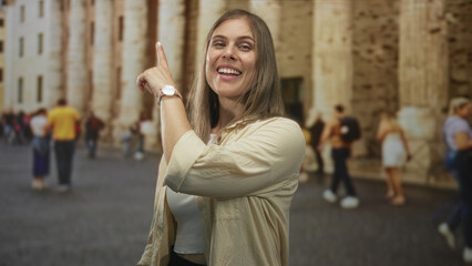 Fototapeta premium Blonde woman wearing a watch on her wrist and pointing finger toward tall stone building on crowded street; excitement.