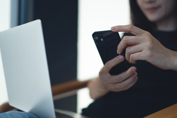 Close up of young smart asian woman using mobile phone, working on laptop computer on her lap at home. Casual business woman online working at coffee shop, freelance at work, online job