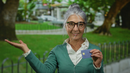 Senior woman in park gestures with hand and holds credit card, showcasing joy outdoors on sunny...