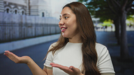 Young smiling woman showing thumb in thumbs up gesture on street lined with fence and trees;...