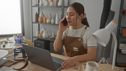Young woman in brown apron holds red phone to ear while typing on silver laptop in studio; productivity.