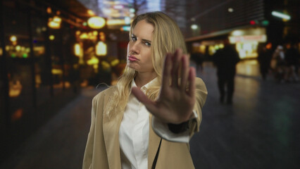 Woman with blonde hair making a stop gesture on a busy city street at night, conveying confidence...