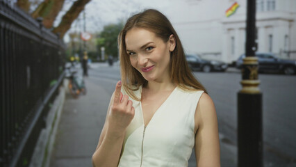 Obraz premium Smiling woman in white dress beckons with finger gesture on busy city street sidewalk near wrought iron fence; invitation.