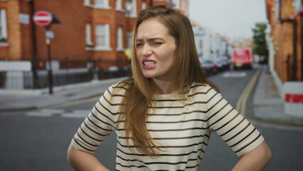 Young woman in striped shirt clutch stomach on urban street in front of brick buildings in daylight; discomfort.
