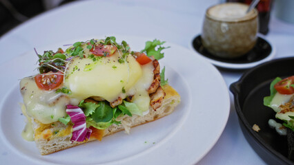 Eggs benedict dish served on a white plate in a cafeteria setting featuring microgreens and tomatoes with a cup of coffee beside.