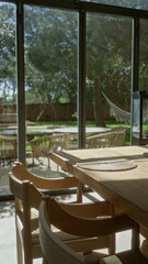 Sunlit restaurant with wooden chairs and table, outdoor view through glass windows, mediterranean decor and hammock visible in garden setting.