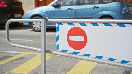 Traffic sign with a red and white no entry symbol stands beside a busy road under a bright sunny sky, with parked cars in the background creating an urban street scene.