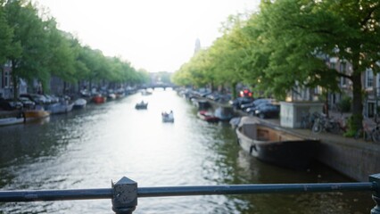 Naklejka premium Blurred canal scene in amsterdam with boats floating on water, trees lining the banks, and defocused cityscape in the background creating a serene dutch atmosphere.