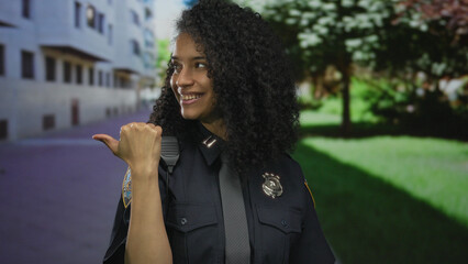 Young hispanic woman police officer in uniform points thumb backward on street with building facade...