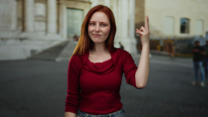 Woman with red hair and red sweater standing on city street looks curious while posing outdoors in urban environment, surrounded by architecture and distant pedestrians.