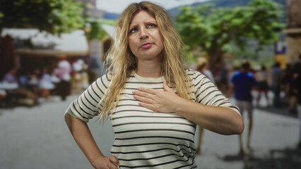 Woman blonde hand on chest in street with blurred crowd and trees, showing discomfort and thought;...