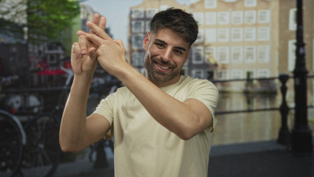 Young hispanic man gestures a hashtag sign with intertwined fingers on a cobblestone street next to canal buildings; connection. - Powered by Adobe