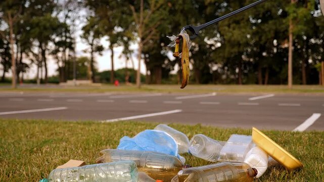A close-up shot of an activist using tongs to collect garbage in an urban area. Plastic garbage and a discarded banana skin are visible.