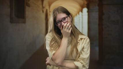 Young blonde woman wearing glasses covers mouth laughing in old town building corridor; joy...