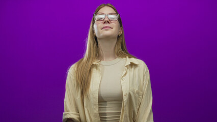 Woman with long blonde hair and subtle smile looks upward wearing glasses and beige shirt in purple studio; confidence self esteem.