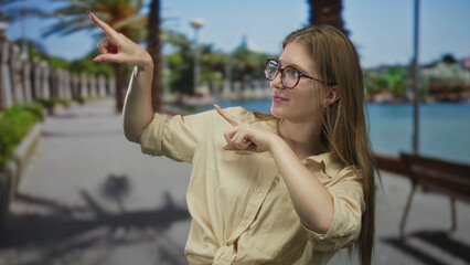 Obraz premium Young blonde woman wearing glasses pointing finger at wooden bench on sunlit street in park; confidence.