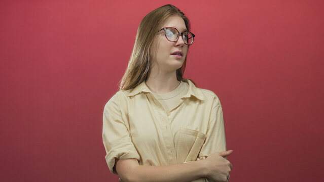 Blonde woman adjusts eyeglasses and rubs temple while wearing beige shirt in red studio setting; stress. - Powered by Adobe