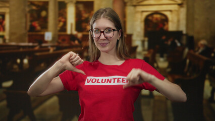 Young blonde woman points finger to volunteer shirt in church building; community service dedication pride.