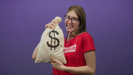 Young blonde woman smiling and holding canvas moneybag with dollar sign in studio wearing volunteer...