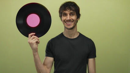 Man holds black vinyl record with pink label and points finger to it in green studio; music promotion excitement.