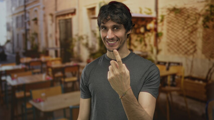 Man pointing finger with smiling wink in sunlit restaurant terrace near wooden tables; playful invitation.