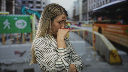 Blonde woman covers mouth with raised hand at street construction site beside yellow barrier and excavator; disgust.