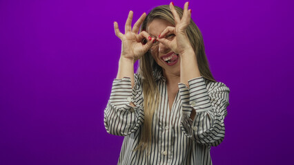 Woman frames her eyes with finger circles making a binoculars sign in a purple studio setting;...