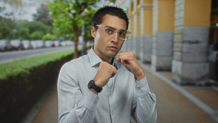 Young hispanic man in glasses practicing boxing stance on urban street, wearing casual shirt, with trees and cars lining the road, conveying focus and determination.
