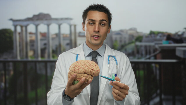 Young hispanic man doctor with stethoscope examines brain model outdoors against roman ruins background.