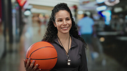 Woman smiles holding basketball inside mall showcasing enthusiasm and energy while wearing sporty...